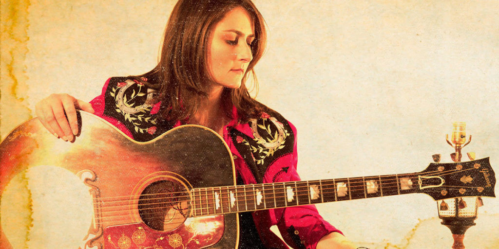 New Country Artist to Watch, Erin Enderlin, holds her guitar while wearing a red and black bedazzled jacket.