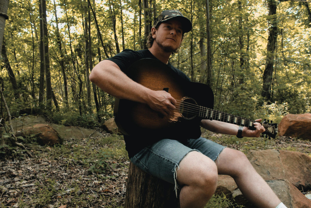 Connor Manley sits on a stump in the woods and plays his guitar