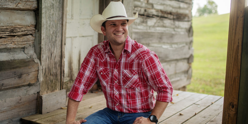 David Adam Byrnes, New Country Artist to Watch, sits on wooden porch while wearing a red flannel shirt, blue jeans, and a white hat.