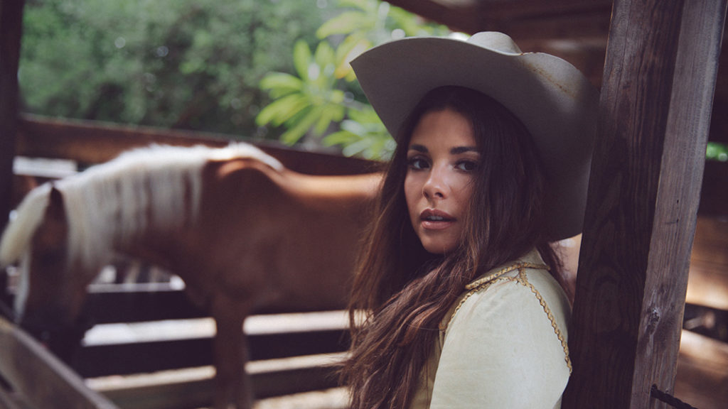 New Country Artist to Watch, Kylie Frey, stands against a wooden post in a horse corral while wearing a white shirt and a grey cowboy hat.